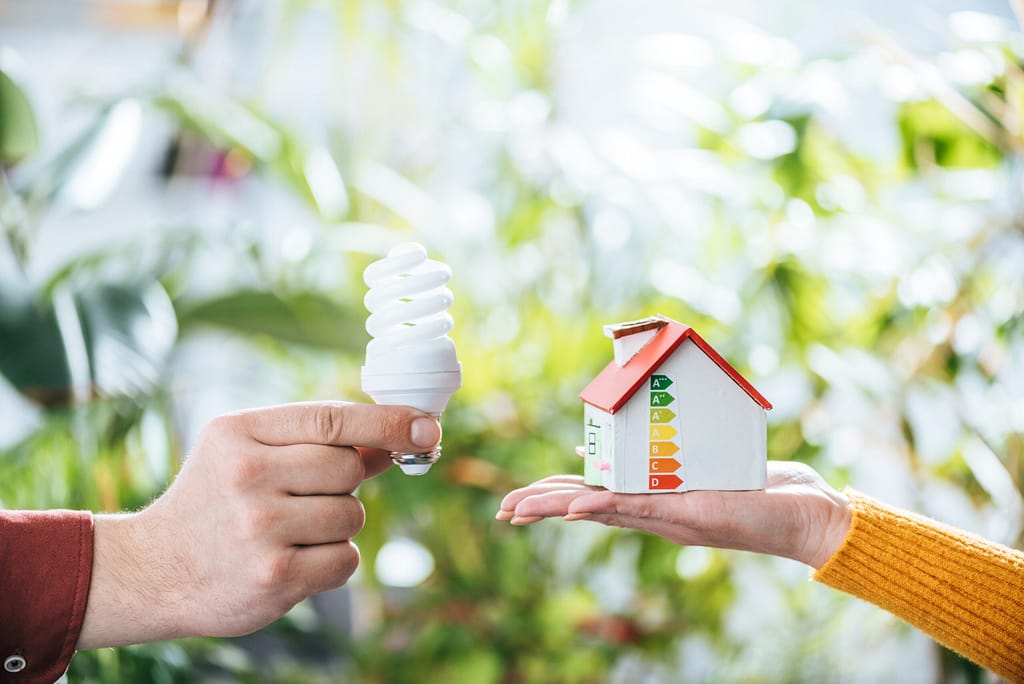 energy efficient homes cost benefit analysis. A hand holding a florescent lightbulb and a toy house representing energy efficiency and cost savings home energy assessments